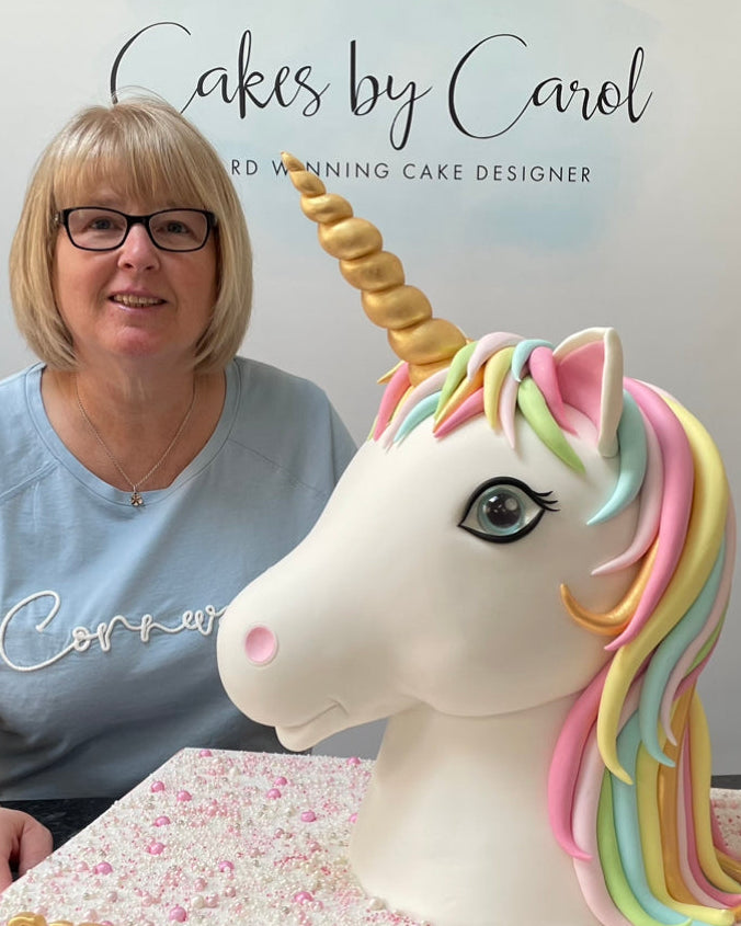 Person sitting next to a unicorn cake with 'Cakes by Carol' sign in the background