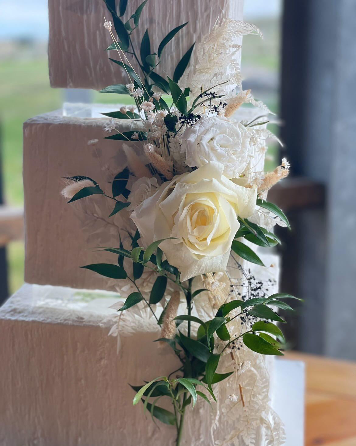 A close up of a square, cream coloured cake with white florals, the layers are separated with 30mm Square Acrylic Cake Separators - Prop Options