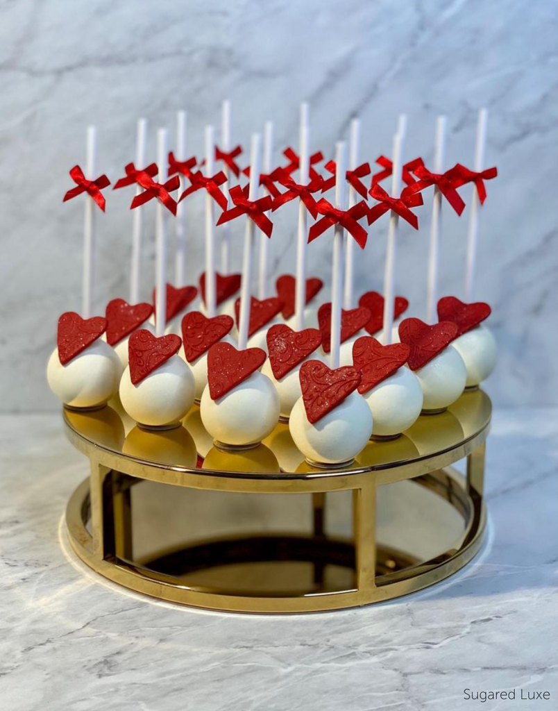 White cake pops with red hearts on them and red bows on the sticks being held on a Gold 10" Round Metallic Cake Spacer - Prop Options