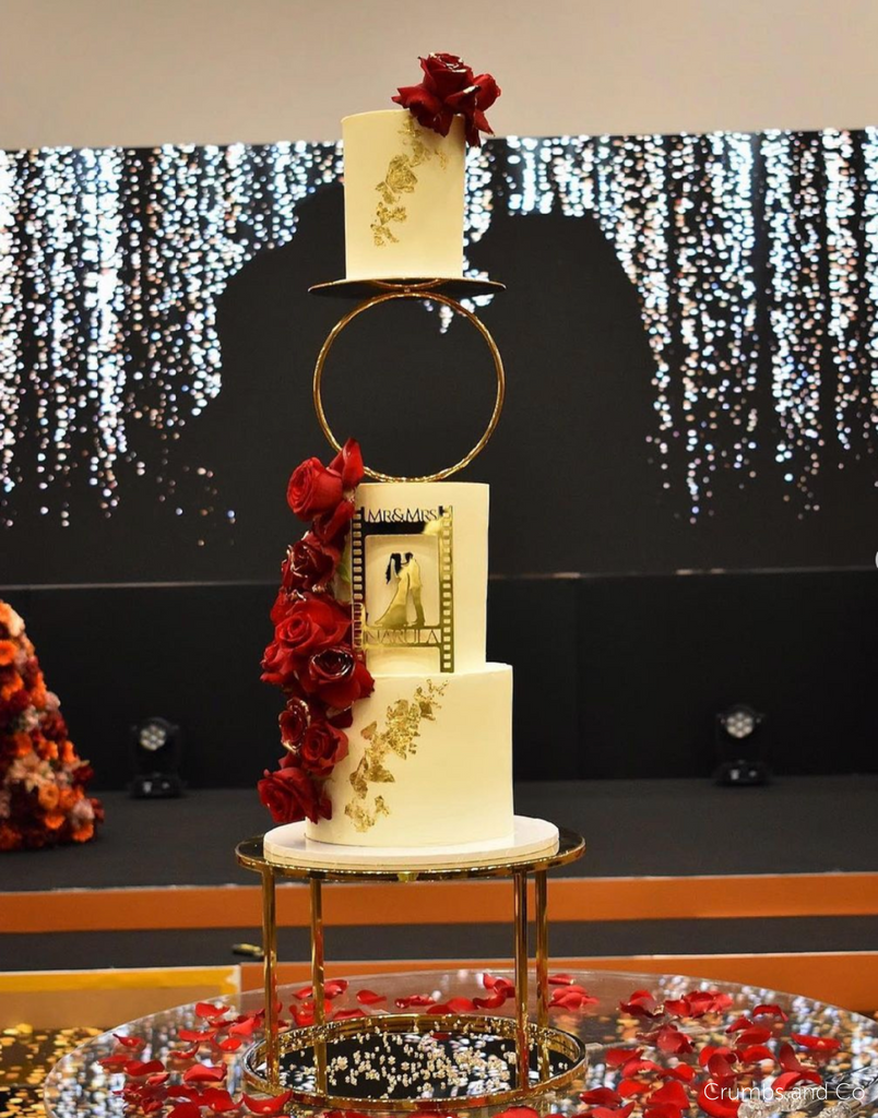 Gold Round Metallic Plinth holding up a white wedding cake with red flowers and gold decorations, the front of the cake has a gold silhouette of the bride and groom and the top layer is held up by a Hoop Spacer - Prop Options