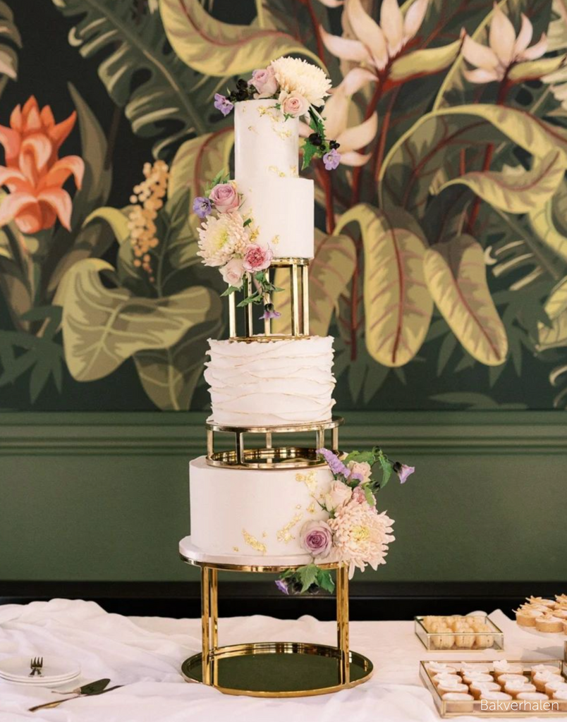 Gold Round Metallic Plinth holding up a white multilayered cake covered in pastel coloured flowers, the tiers are separated by gold spacers - Prop Options