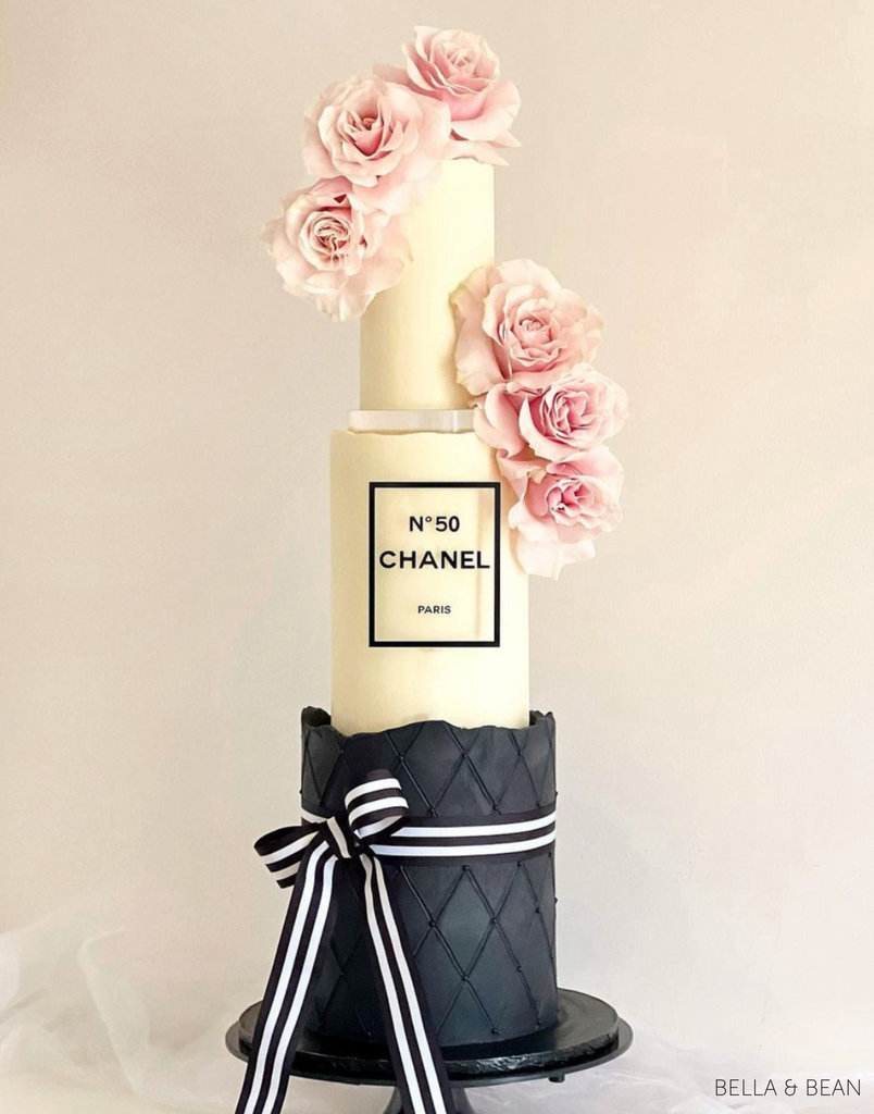 A three tiered cake with a black bottom tier with a white and black ribbon and a cream top and middle tier with a No 50 Chanel Paris logo and pink rose decorations, the top two layers are separated with a 15mm Acrylic Cake Separator - Prop Options