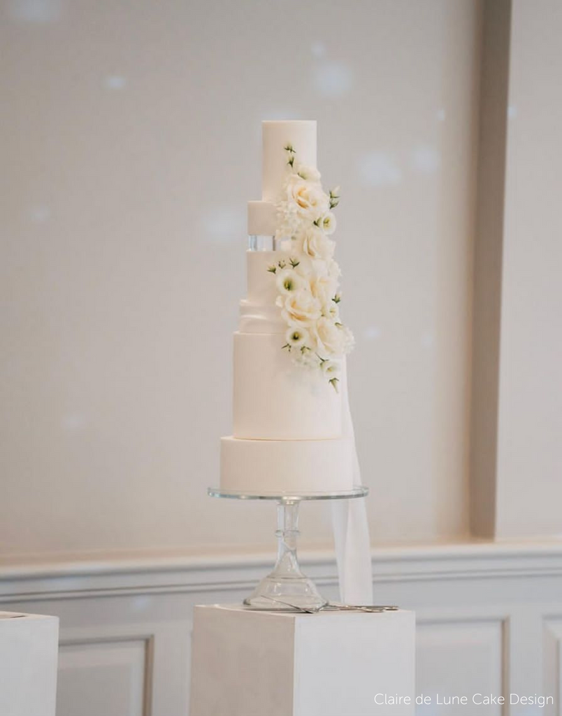 A simple white multitiered cake with white flowers along the side, two of the tiers are separated by a 30mm Round Acrylic Cake Separator - Prop Options