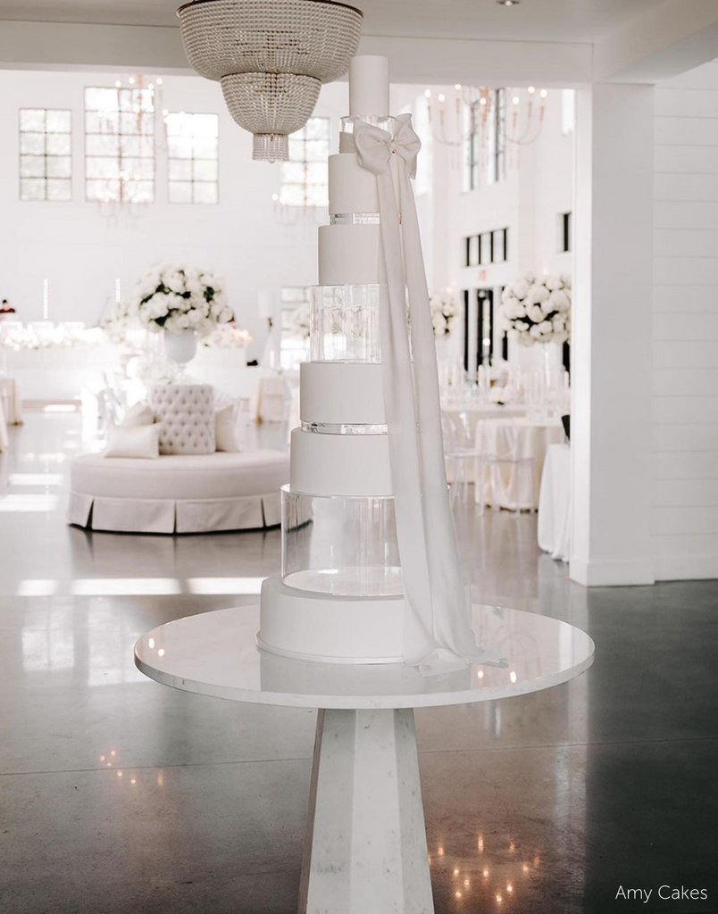 A plain white multitiered cake with a long white ribbon, each of the tiers is separated alternating between a clear tier and a 30mm Round Acrylic Cake Separator - Prop Options