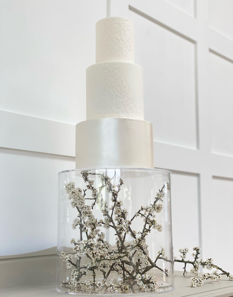 Round Acrylic Fillable Plinth full of flower branches and holding a multilayered white cake - Prop Options