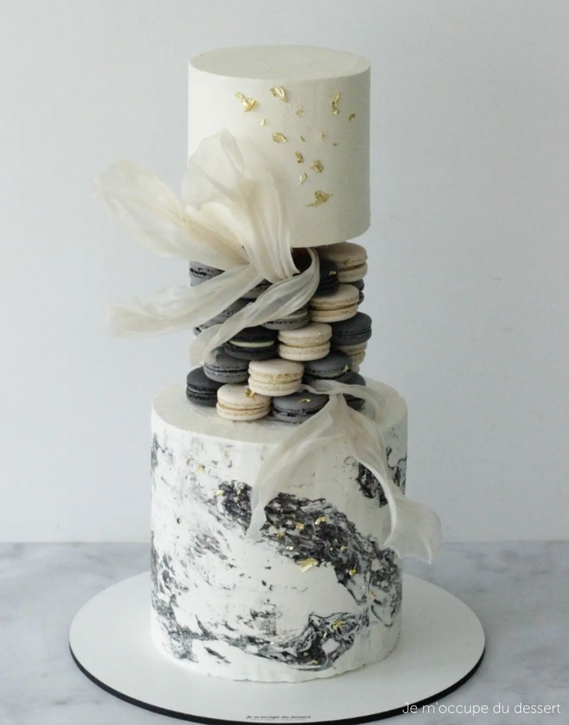 A two tier white cake with black and gold marbled decorations, between the two tiers there are neatly stacked black and white macarons resting on a PropSecure® Adjustable Central Bar Cake Separator - Prop Options