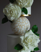 A close up of two plain white cake tiers covered in white flowers with a white and gold ball between them resting on a PropSecure® Adjustable Central Bar Cake Separator - Prop Options