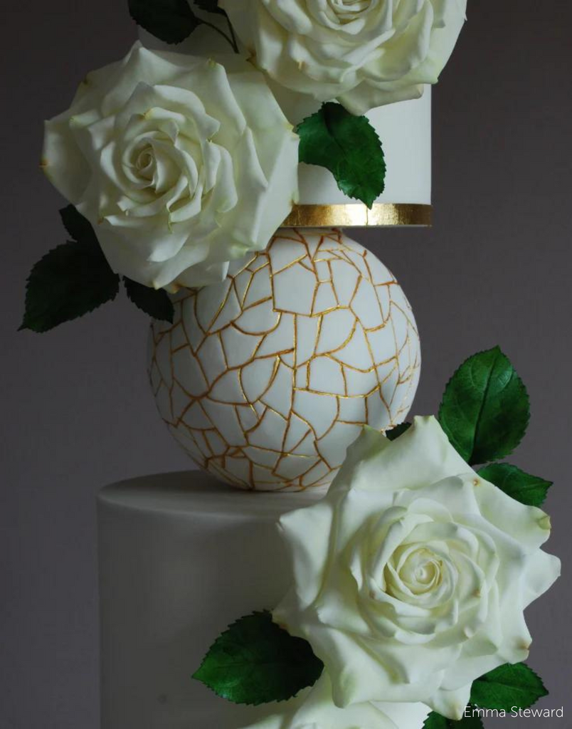 A close up of two plain white cake tiers covered in white flowers with a white and gold ball between them resting on a PropSecure® Adjustable Central Bar Cake Separator - Prop Options