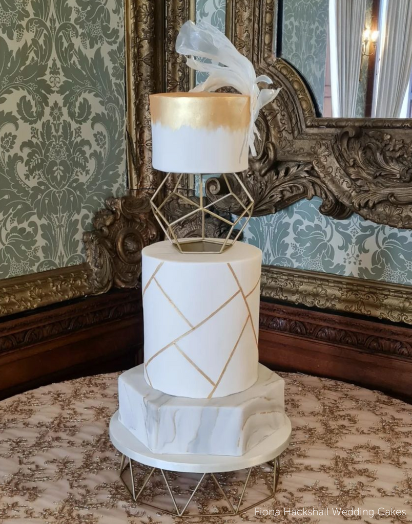 A white three layer cake with subtle gold and grey marbing on the layers, the top two layers are separated by a Geometric Pentagon Spacer - Prop Options