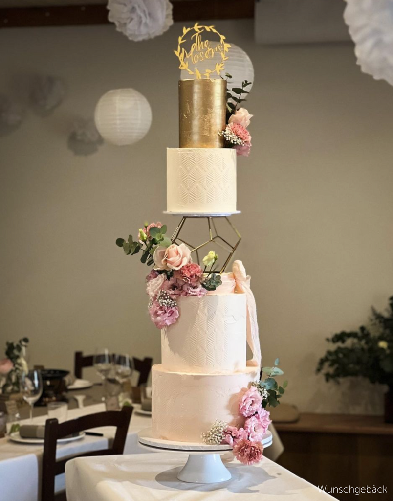 A four layer cake with three plain white tiers and a gold tier on top, there are pink flowers on each of the white tiers and a gold topper, the middle of the cake is separated by a Geometric Pentagon Spacer - Prop Options