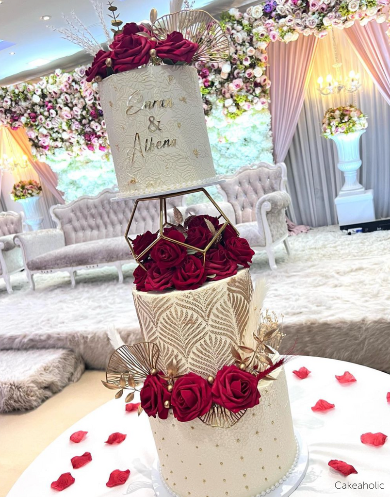 A white three tier cake with elegant gold patterns on each layer, dark red roses, and the names Elena & Athena in gold, the top two layers are separated by a Geometric Pentagon Spacer - Prop Options