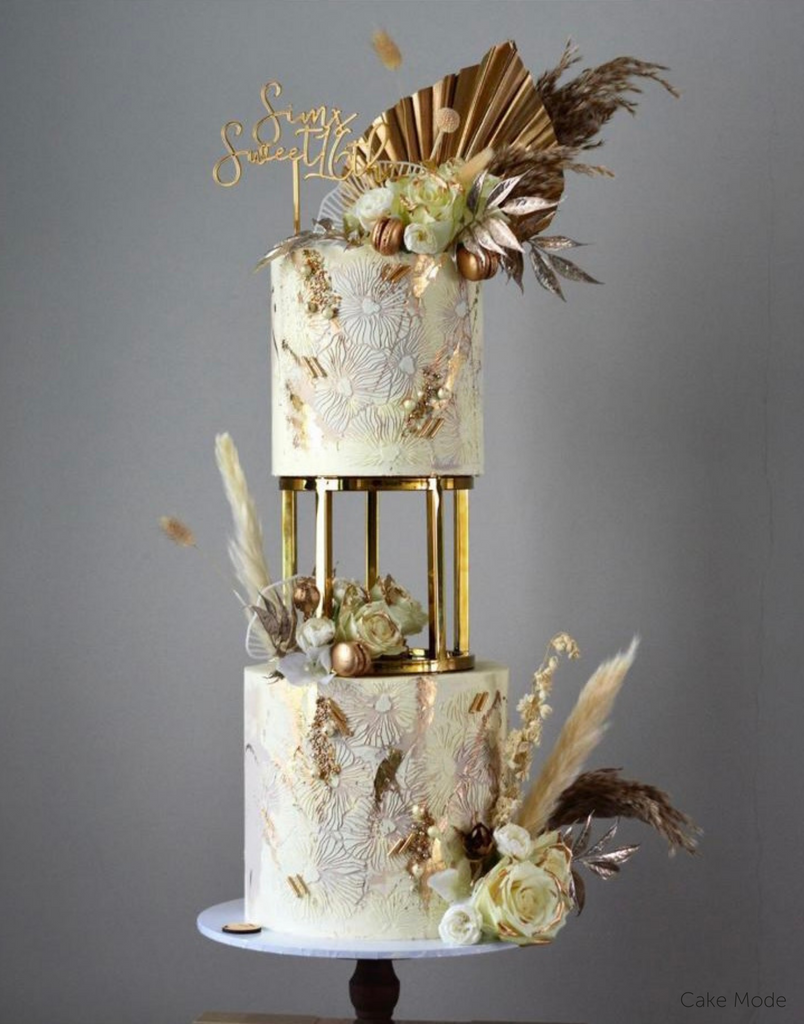 A white two tier cake with gold floral designs and various light coloured flowers on the top and bottom, the tiers are separated by a 6" Round Metallic Cake Spacer - Prop Options