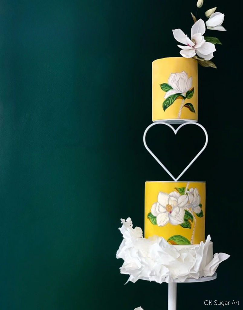 A bright yellow two tier cake with white flowers painted on the front and real white flowers on the top, the layers are separated by a Heart Tier Separator - Prop Options