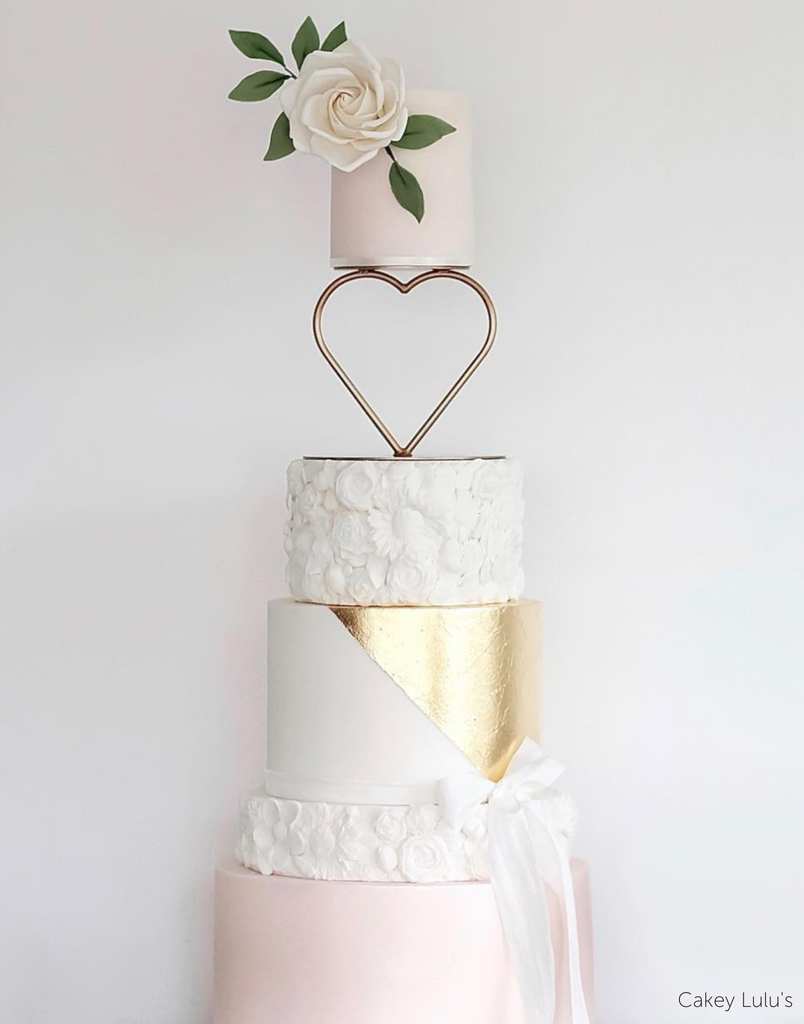 A white and light pink multitiered cake with subtle floral piping on two layers, a white ribbon, and a white rose on the top, the top two layers are separated by a Heart Tier Separator - Prop Options