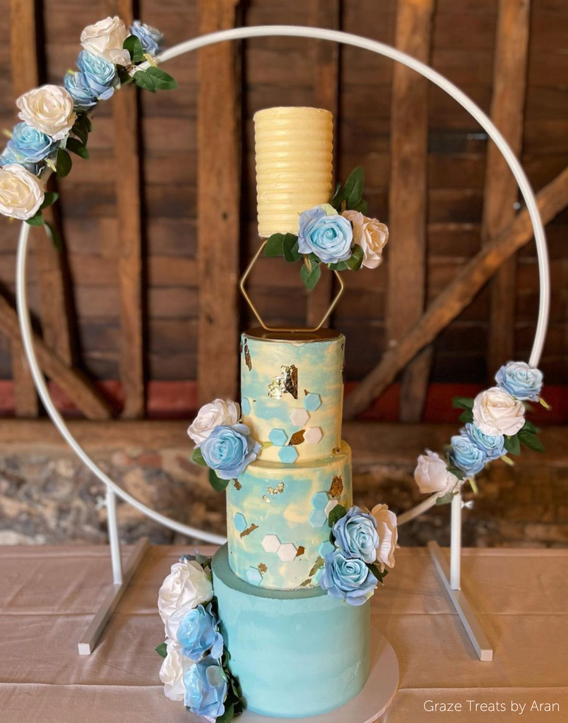 A mixed blue and cream four layer cake with blue and cream flower decorations on a large hoop, the top two tiers are separated by a Hexagon Tier Separator - Prop Options