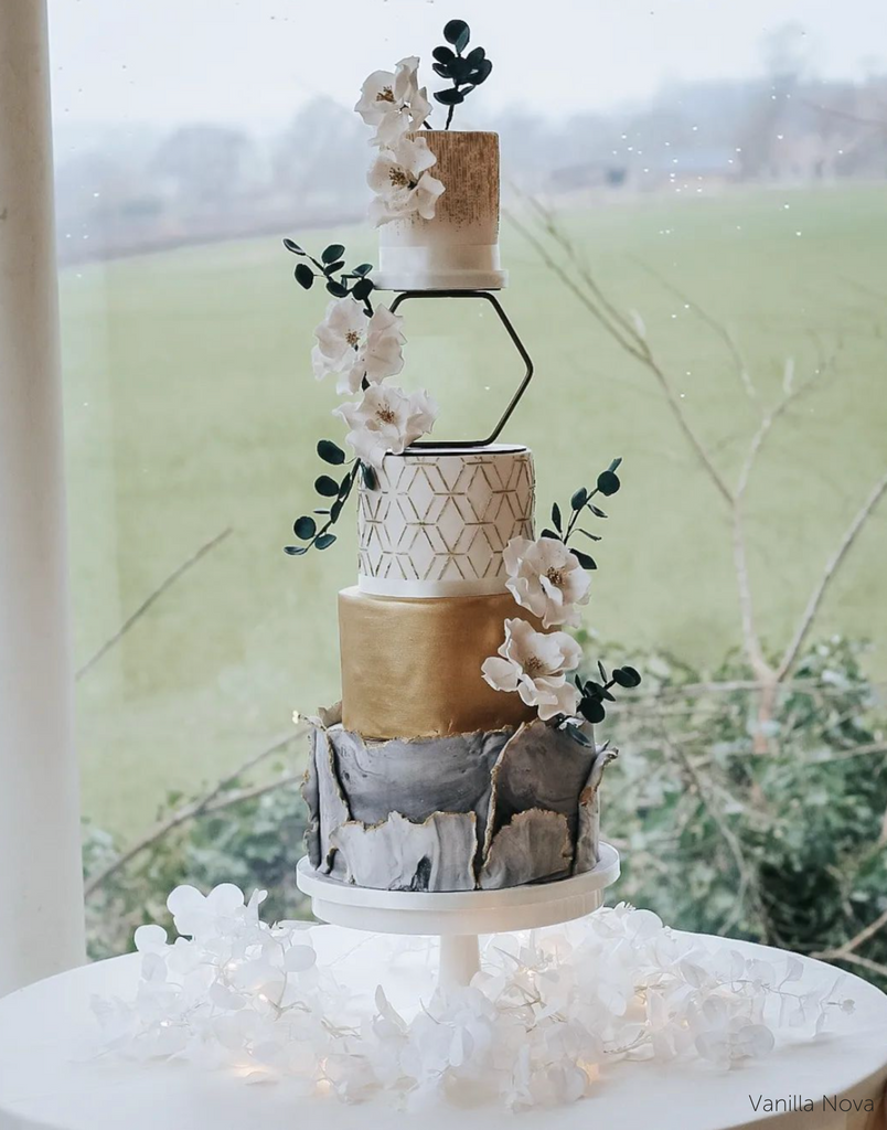 A four tier cake with grey, gold, white, and brown layers covered in white flowers, the top two layers are separated by a Hexagon Tier Separator - Prop Options