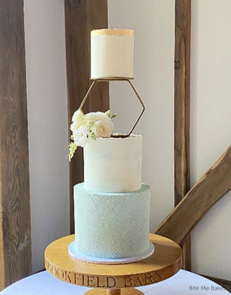 A three tier cake with two plain white tiers and a light blue bottom tier with a scallop pattern, the top two tiers are separated by a Hexagon Tier Separator with white roses on it - Prop Options