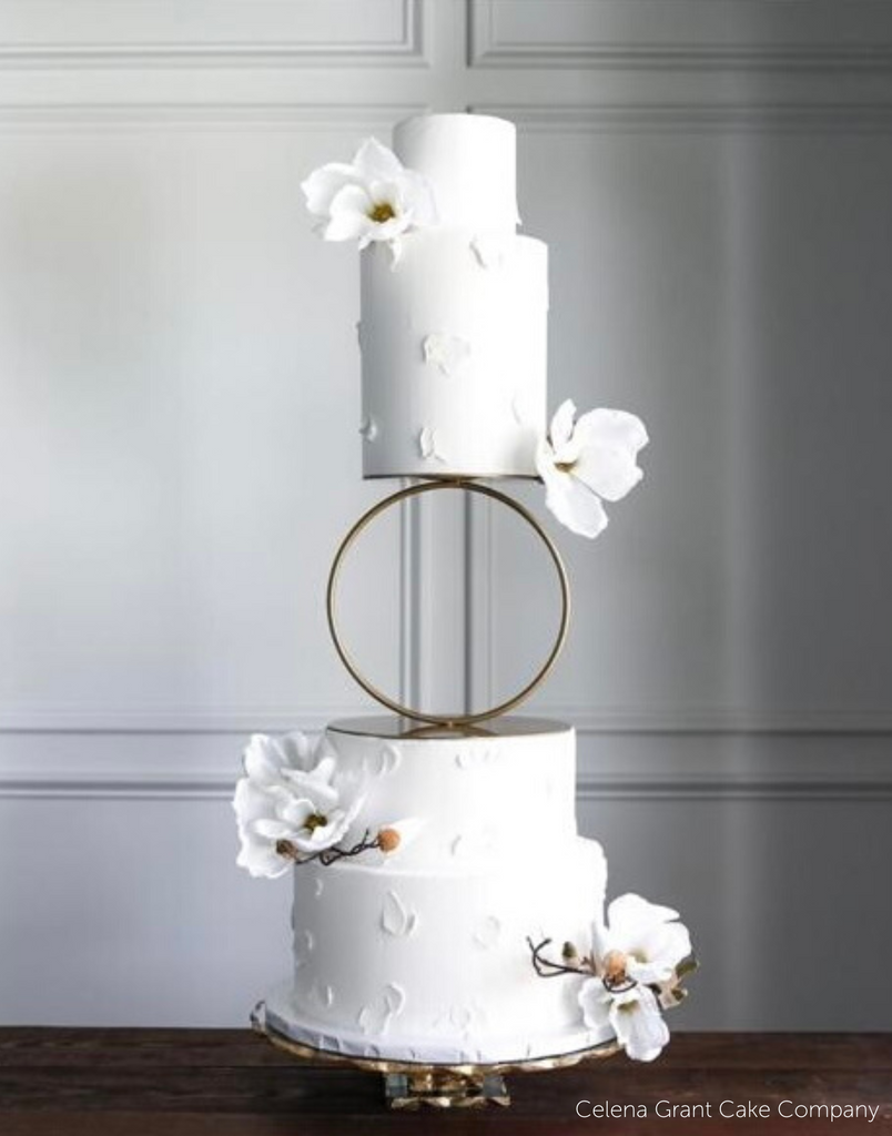 A four tier white cake with subtle floral patterns and white flowers attached to each tier, the middle of the cake is separated using a Hoop Tier Separator - Prop Options