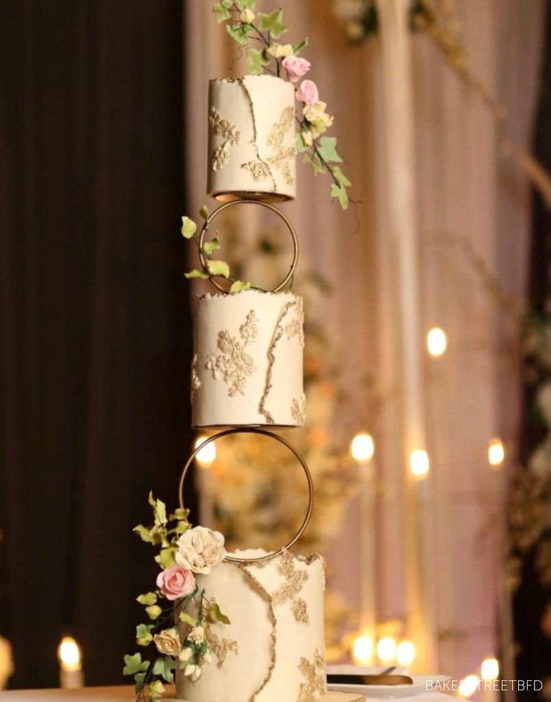 A tall white cake with subtle gold decorations and pastel coloured flowers, each tier is separated by a Hoop Tier Separator - Prop Options