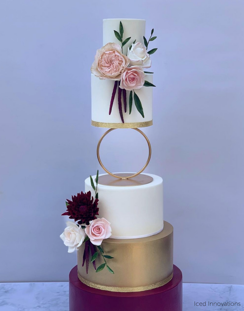 A five layer cake, the first three layers are white, the fourth layer is gold, the fifth layer is purple, there are purple and pink flowers along the top and bottom, the middle of the cake is separated using a Hoop Tier Separator - Prop Options