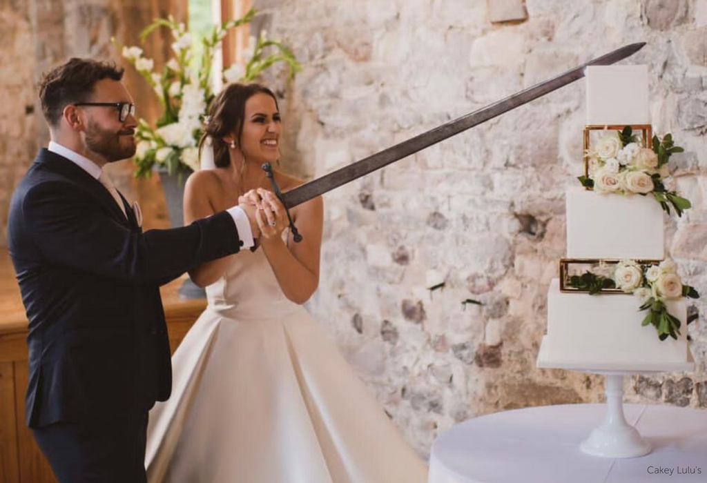 A couple cutting into a wedding cake with a sword, the cake is white with white roses and is separated using Square and Rectangle Metallic Cake Spacers - Prop Options
