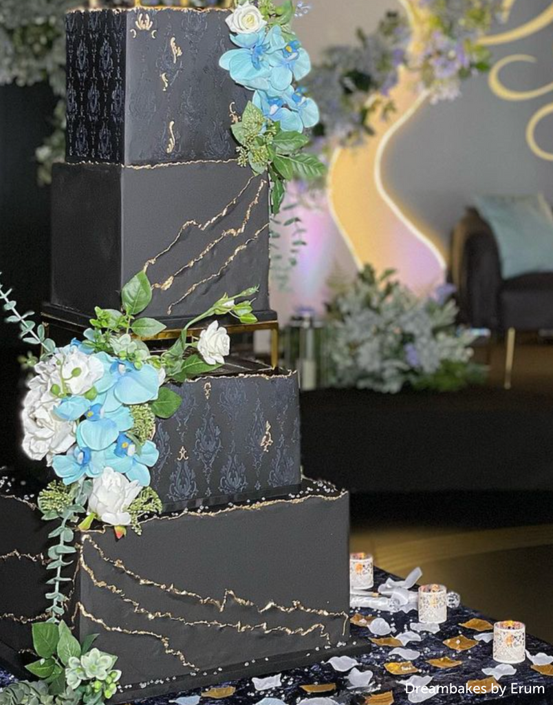 A black multilayered cake with subtle gold accents and blue flowers, the middle layers are separated by a Rectangle Metallic Cake Spacer - Prop Options
