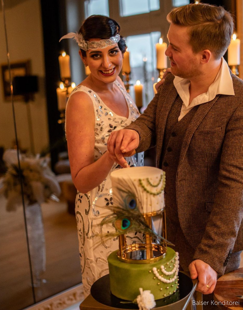 A couple in Roaring Twenties outfits cutting a green and white cake decorated with a peacock feather, the cake tiers are separated using a 6" Round Metallic Cake Spacer - Prop Options