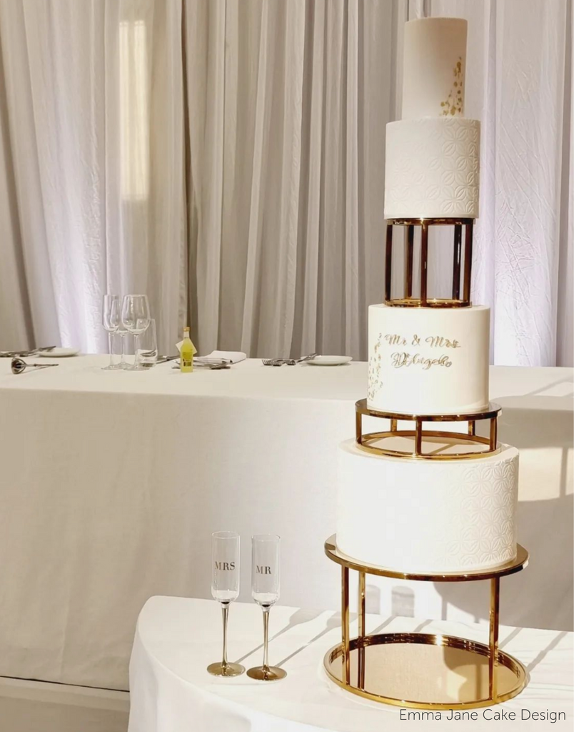 A plain white four layer wedding cake with gold lettering, the tiers are separated using 6" & 10" Round Metallic Cake Spacers - Prop Options