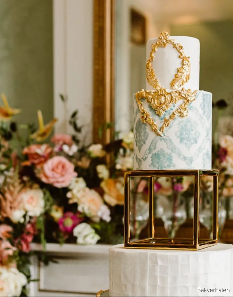A white three layer cake with light blue and gold accents, the bottom tiers are separated using a Square Metallic Cake Spacer - Prop Options