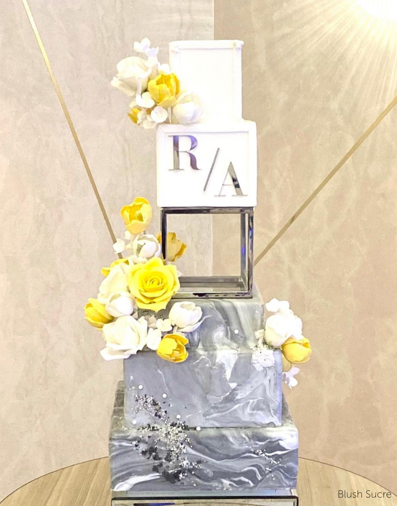 A four tier grey and white cake with silver lettering and yellow flowers, the middle of the cake is separated using a Square Metallic Cake Spacer - Prop Options