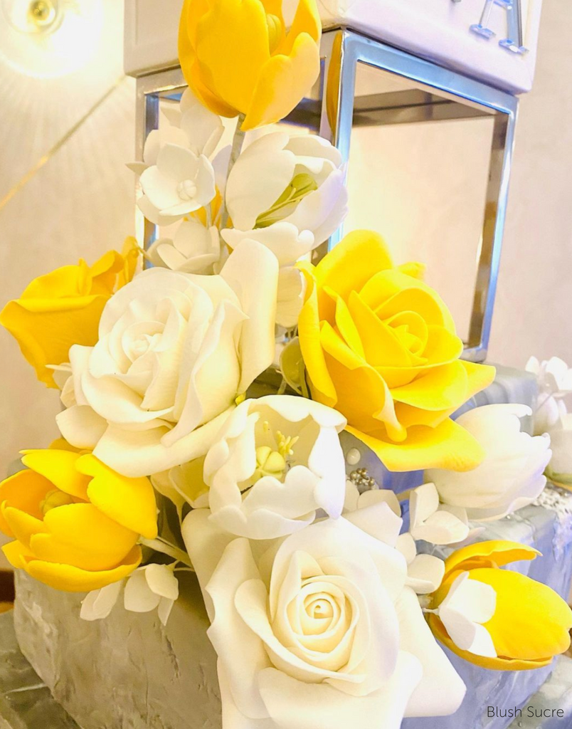 A close up of a cake using a Square Metallic Cake Spacer covered in white and yellow flowers - Prop Options