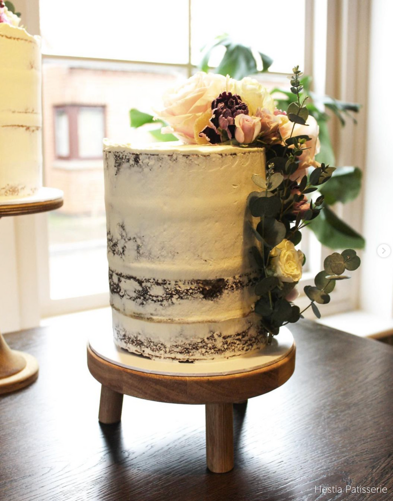 A rustic cake covered in flowers stood on a Solid Oak Tripod Cake Stand - Prop Options
