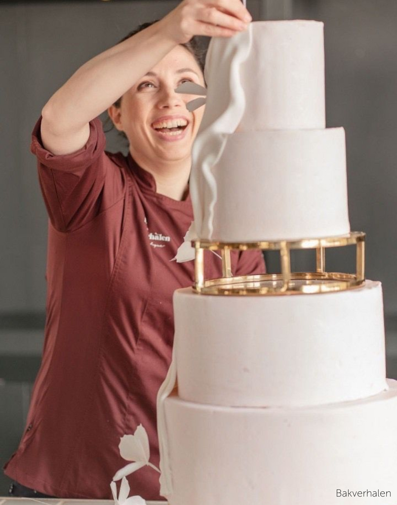 Baker adding decorations to a multitiered white cake, in the middle of the tiers is a Gold 10" Round Metallic Cake Spacer - Prop Options