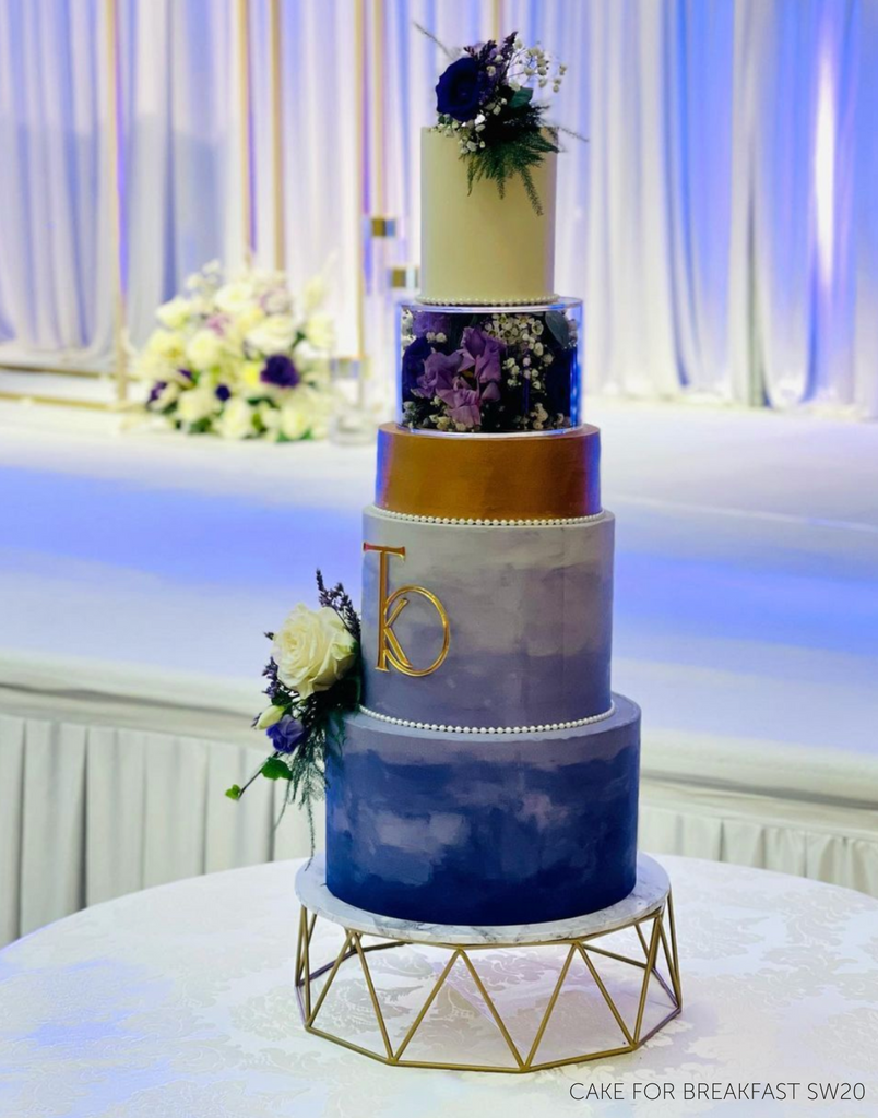 Geometric Cake Stand holding a five layer cake with blue, purple, gold, and white layers with floral decorations and gold lettering on the front - Prop Options