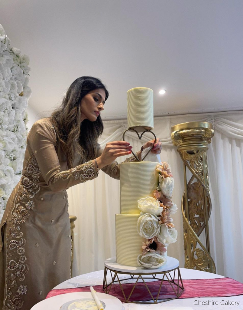 Geometric Cake Stand holding a white three layer cake with white and peach flowers along the side as a woman adjusts a heart spacer between the top two layers - Prop Options