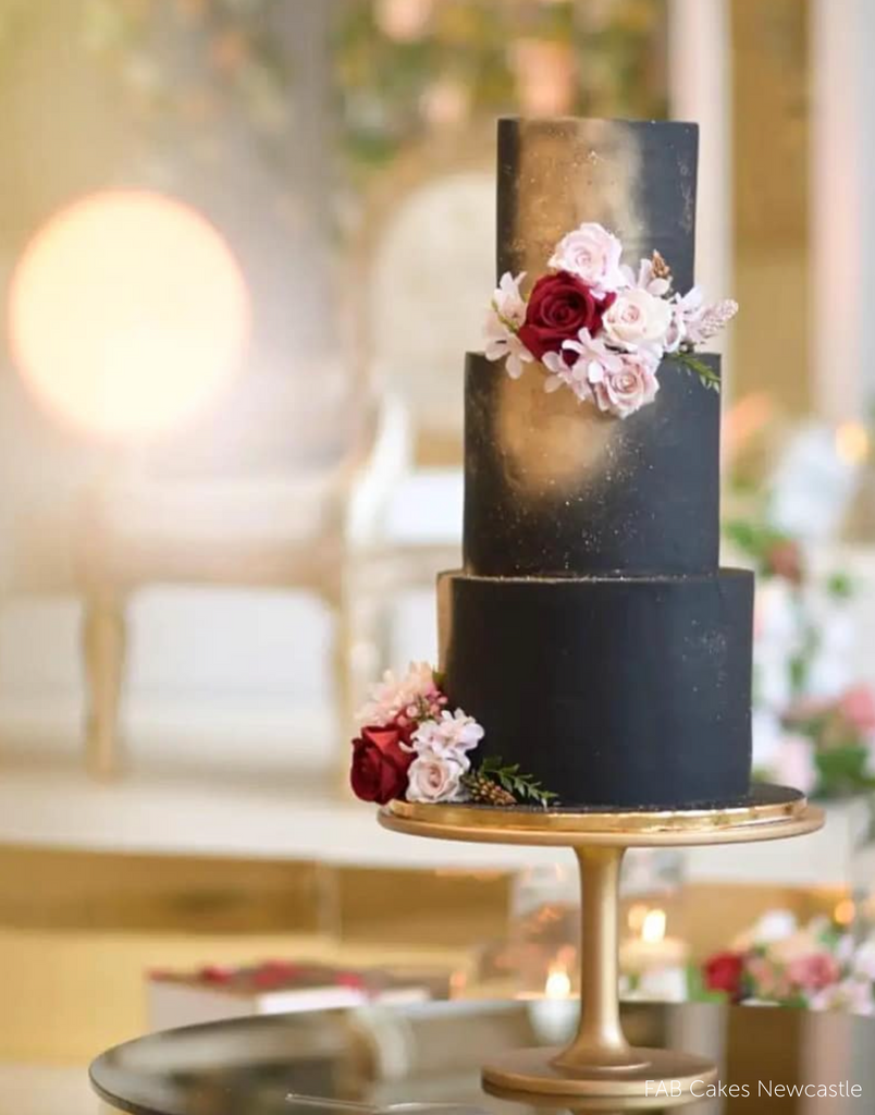 A black cake covered in red and pink flowers stood on The Hourglass Scandinavian Birch Cake Stand - Prop Options