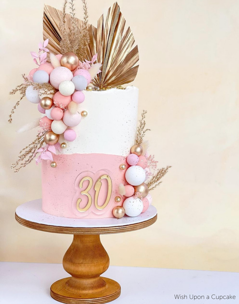 A white and pink cake with pastel coloured ball decorations and the number 30 in gold lettering sat on The Teardrop Scandinavian Birch Cake Stand - Prop Options