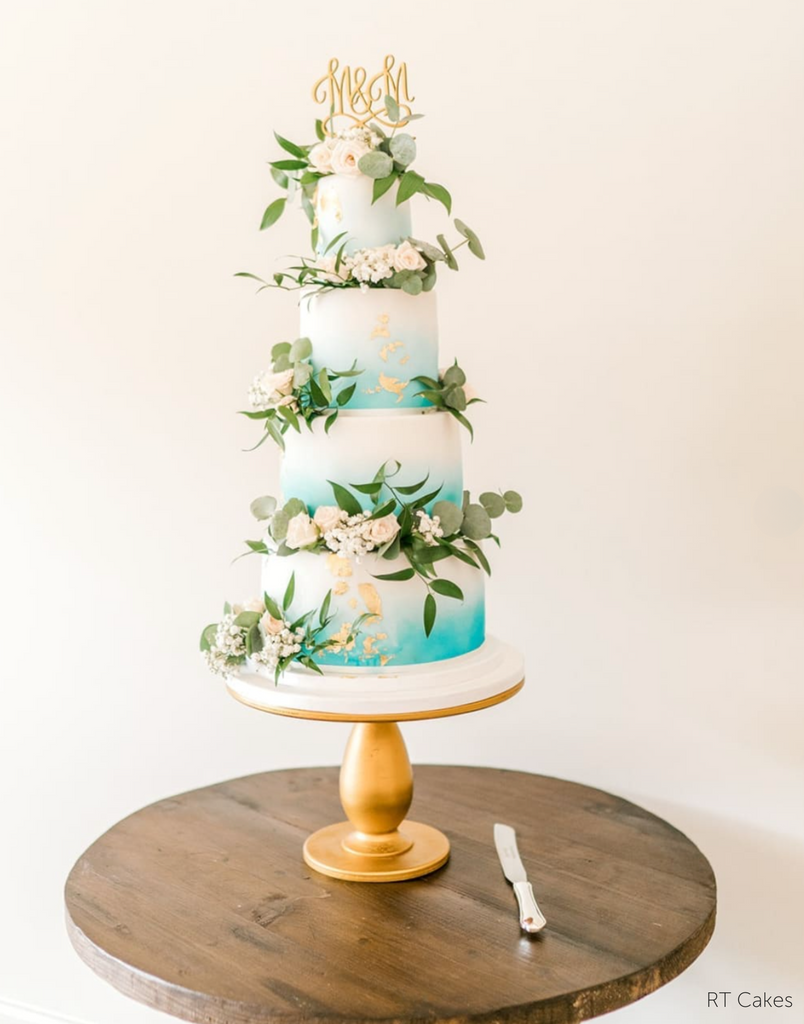 A white and blue cake with gold accents and white flowers stood on The Teardrop Scandinavian Birch Cake Stand - Prop Options