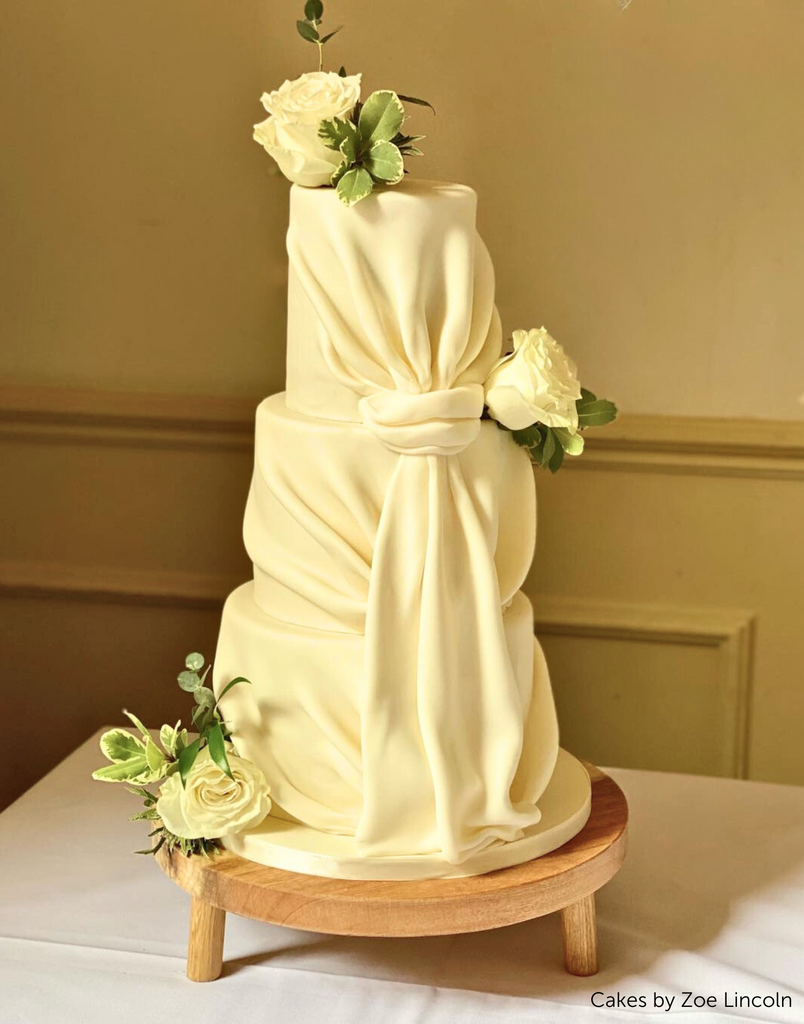 A tall cake covered in cream coloured fondant and white roses on top of a Solid Oak Tripod Cake Stand - Prop Options
