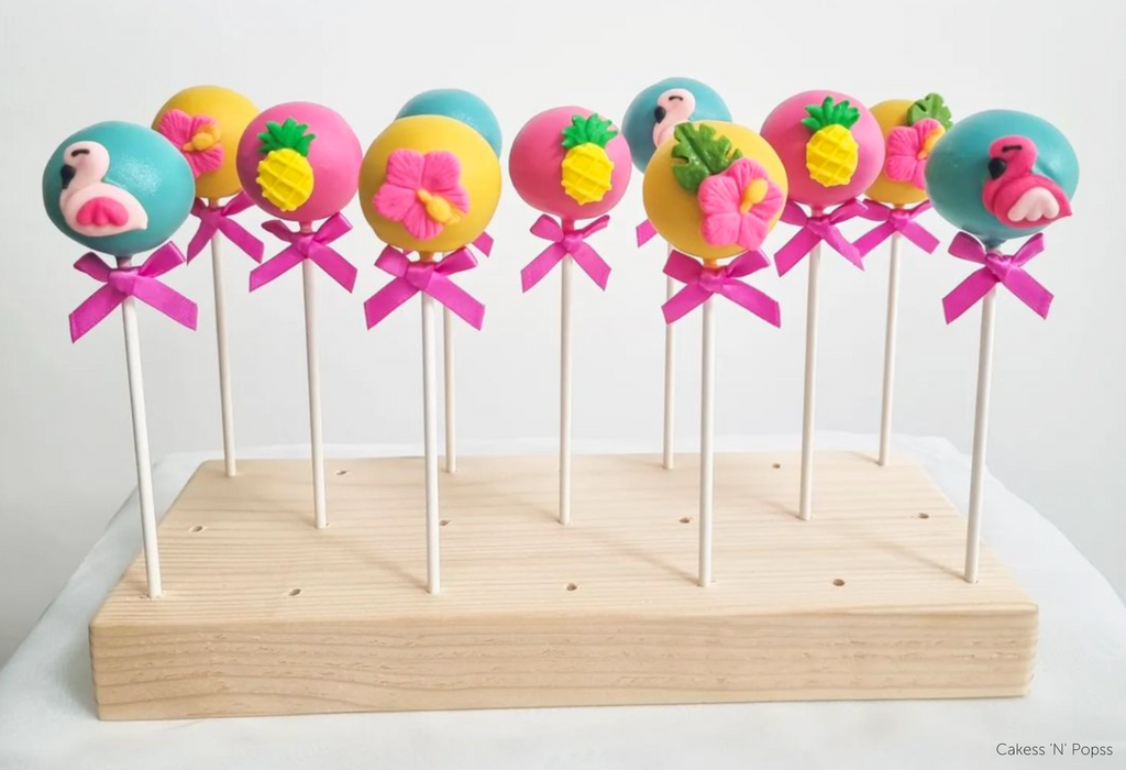 Tropical themed cake pops in yellow, pink, and blue with various flamingo, flower, and pineapple designs and finished with a pink bow on each stick, they're stood inside an English Pine Wooden Cake Pop Stand - Prop Options