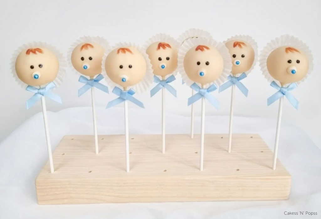Cake pops decorated to look like baby boys in white bonnets with blue bows on each stick held inside an English Pine Wooden Cake Pop Stand - Prop Options