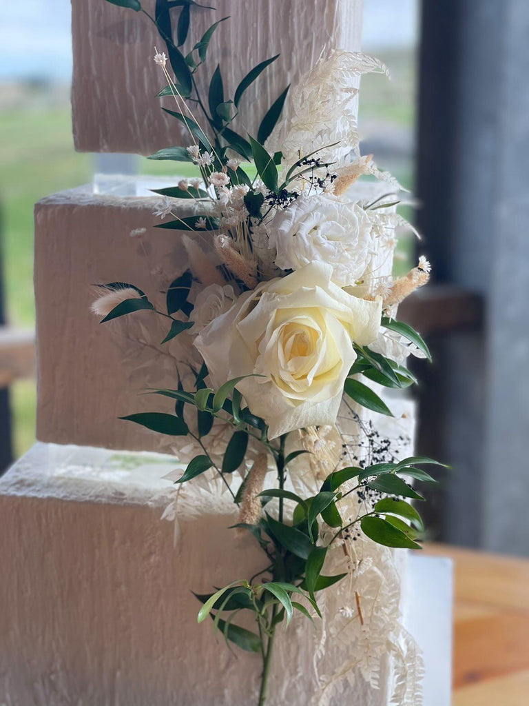 A close up of a square, cream coloured cake with white florals, the layers are separated with 30mm Square Acrylic Cake Separators - Prop Options